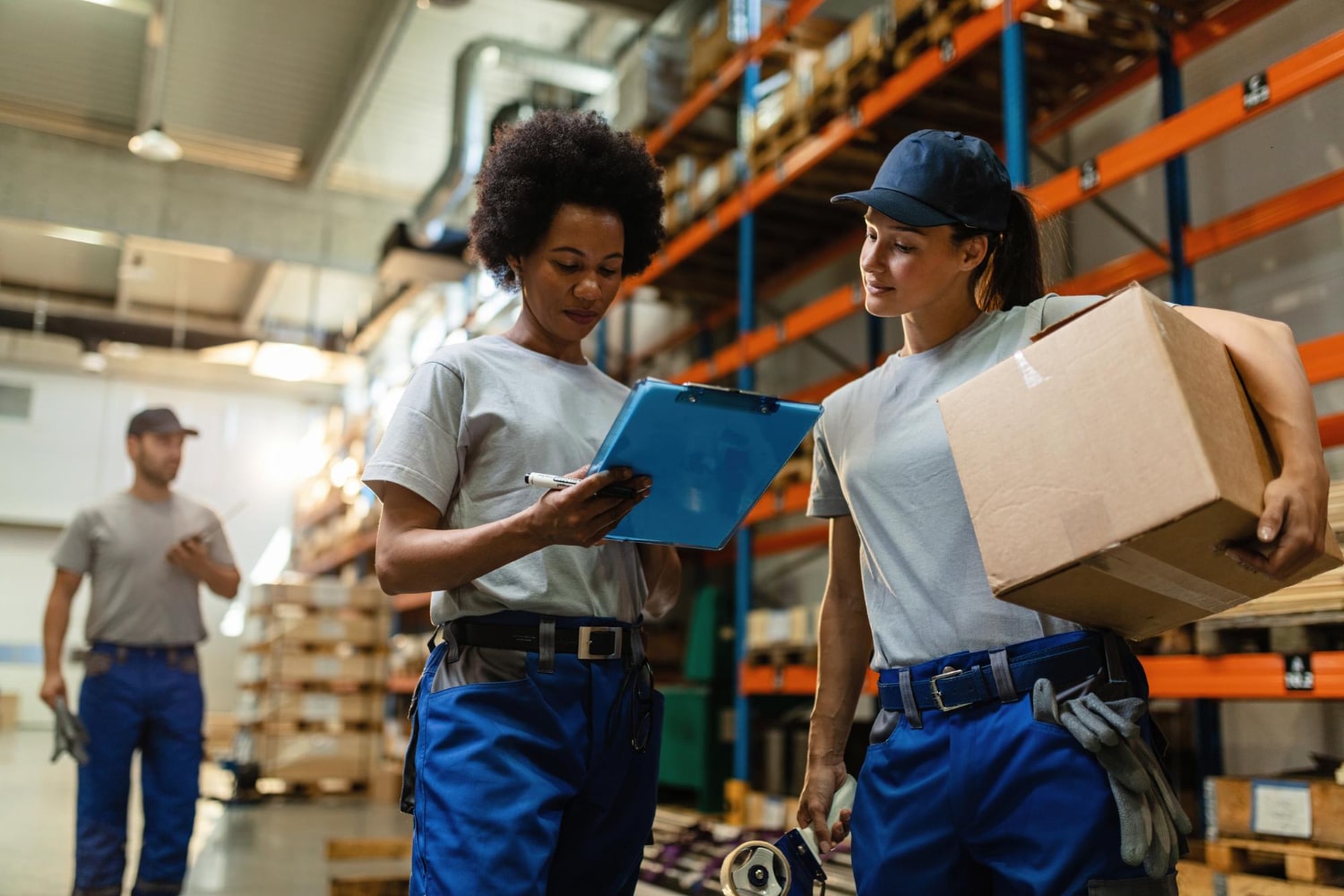 Two women working in a warehouse look at a clipboard, the woman on the right has a box under her arm